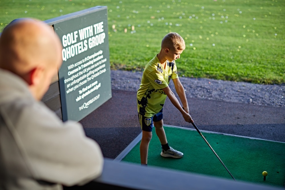 A young boy in a yellow top on the golf driving range lining up to hit the golf ball with jis junior golf club whilst an adult professional golfer guides.